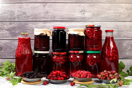 Jars with raspberry, cherry, gooseberry jam and compote on kitchen background with leaves and fresh berries, home canning concept, banner and advertisement of shop and market, village harvest, selective focusの写真素材