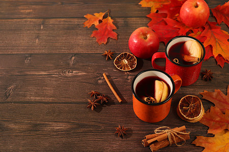 Two colorful enamel mugs with hot autumn drink, mulled wine with apples and autumn fallen leaves, star anise and cinnamon on a wooden table, banner for display, autumn greeting concept, mockup for menu.の写真素材