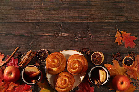 Cinnamon rolls with hot autumn drink, mulled wine with apples and autumn fallen leaves, star anise, ginger and cinnamon on wooden table, banner for display, autumn greeting concept, mockup for menu.の写真素材