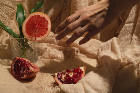 The composition is made on a curtain with grapefruit and pomegranate and a glass full of plants. There is a hand that wants to take the glass.の写真素材
