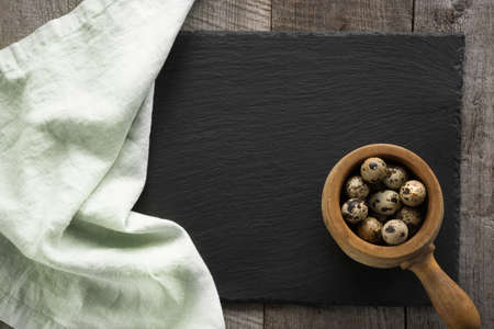 Green linen napkin on black slate dish and vintage wooden bow with quail eggs for cooking on wooden background. Space for your text.の写真素材