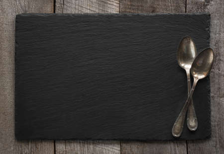 Black slate plate and vintage spoons on wooden board. Top view. Space for your text.の写真素材