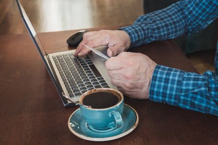 Side view shot of a man's hands holding card and using laptop sitting at wooden table in office with cup of black coffee.  Close up.の写真素材