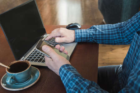 Side view shot of a man's hands using smart phone and laptop sitting at wooden table with cup of black coffee in interior.  Close up.の写真素材