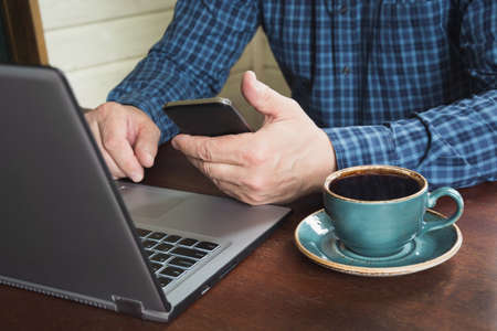 Side view shot of a man's hands using smart phone and laptop sitting at wooden table with cup of...