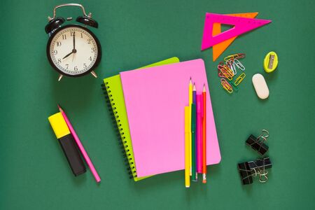 Colorful school supplies, pink book, and alarm clock on green background. Top view, flat lay, copy...