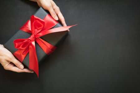 Gift box wrapped in black paper with red ribbon in female hand on black surface. Copy space. Top view.の写真素材
