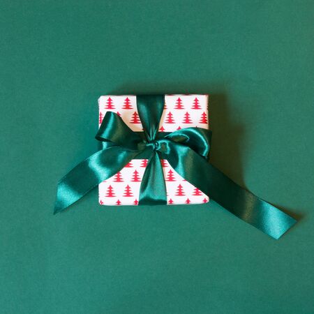 Gift box wrapped in design paper with green ribbon on green surface. Top view. Square image.