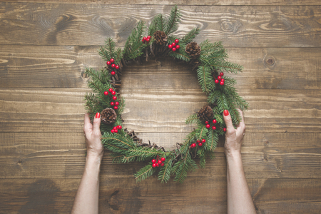 Christmas wreath made of spruce branches with holly berries on wooden board. Female hand holding wreath. Top view.の写真素材