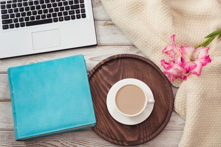 Female workplace with a laptop, cup of coffee, blue notebook, cozy sweater  and pink flowers on light wooden background.Top view composition with copy space.の写真素材