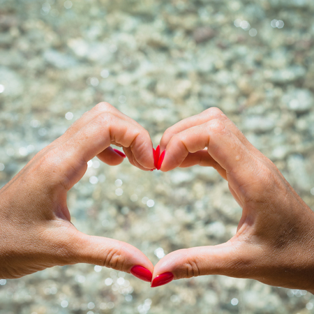 Female hand with red nail in shape of heart, beach as background. Love summer concept.の写真素材