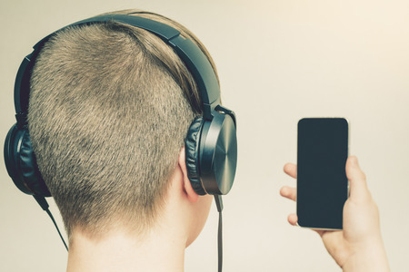 Young blond guy listening to music by headphones and hold smartphone on grey background. Isolated. Close up.の写真素材