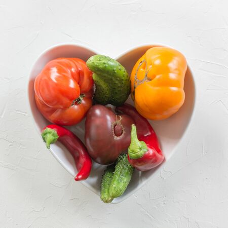 Oddly shaped organic colorful tomato, pepper, cucumber in dish as heart on white concrete. Concept of organic vegetables. View from above.の写真素材