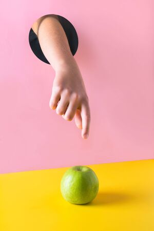 Childs hand through hole in pink paper reaches for green apple on yellow background. Food concept.の写真素材
