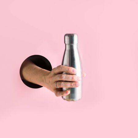 Metal reusable bottle in female hand through in pink hole. Trendy concept.の写真素材