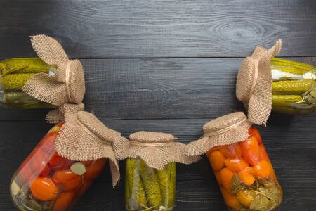 Canned vegetables cucumber and tomato in glass jars on wooden dark board. Space for text. View from...