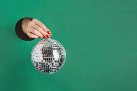 Female hand holding mirror disco ball through round hole in green paper. Invitation to Christmas...