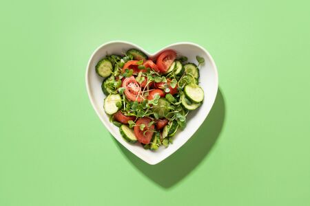 Fresh salad with tomato, cucumber, vegetables, microgreen radishes in plate shape of heart on green. View from above. Concept vegan and healthy eating.の写真素材