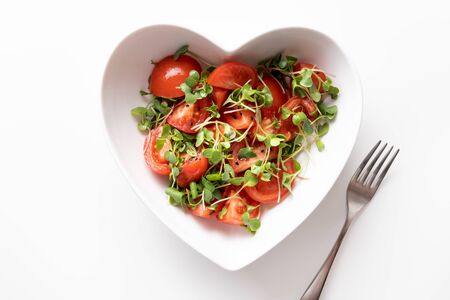 Fresh salad with tomato and microgreen radishes in diah shaped of heart isolated on white. Top view. Concept vegan and healthy eating. Isolated on white.の写真素材