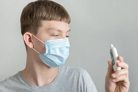 Teenager in medical mask holds thermometer in his hands and measures temperature. Covid 19....