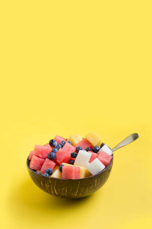 Fruits salad of melon, watermelon, blueberry in coconut bowl on yellow background. Healthy breakfas. Clean eating. Vertical. Negative space.の写真素材