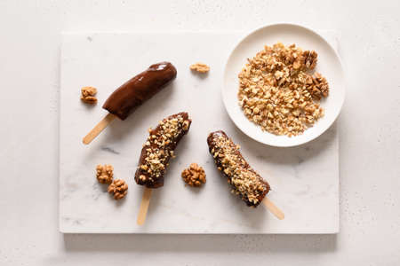 Frozen banana lollipops or lollies with walnuts sprinkles for kids on white. Easy vegan sweets. View from above.の写真素材