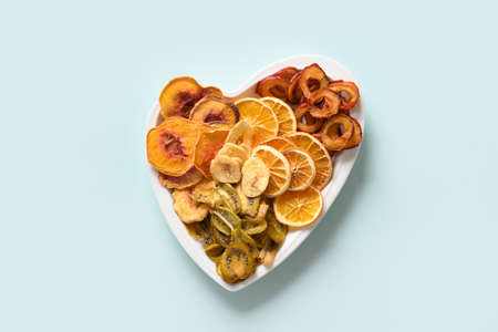 Dried sliced plums, kiwi, peach fruits chips in plate shape of heart on blue background, home drying. Snack vegan free sugar food. View from above.の写真素材