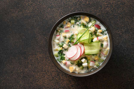 Traditional Russian cold vegetables okroshka soup with chopped cucumber, radish, potato, boiled eggs and kefir or airan, carbonated water isolated on a brown background. Top view.の写真素材