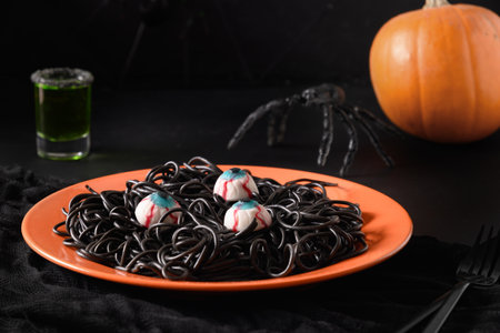 Halloween Italian black pasta squid ink decorated spiders, eyes in orange plate with potion on black table. Horror funny food for party. Close up.の写真素材