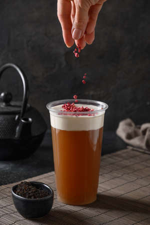 Woman sprinkles Chinese cheese tea in plastic glass with whipped cream cheese topped berrires on black background. Vertical format.の写真素材