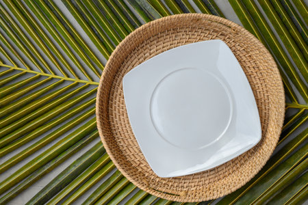 Empty square white plate in authentic wicker tray on green palm leaf. View from above. Space for design or text. Template for Asian and tropical cuisine.の写真素材