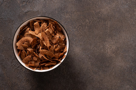 Coconut chips with cinnamon in white bowl on brown background. Tasty sugar free vegan snack. View...