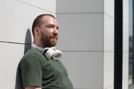 Portrait of caucasian handsome beard man wearing military color t-shirt with headphones near modern...