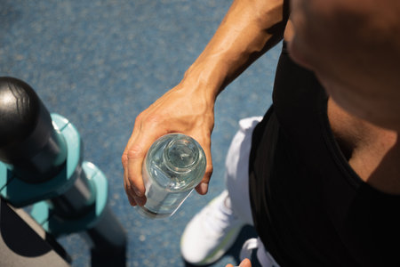 Athletes man holding glass bottle of water on outdoor sports ground. Concept water balance for hot season for active workout. Close up. View from above.の写真素材