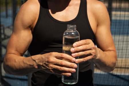 Athletes man holding glass bottle of water on outdoor sports ground. Concept water balance for hot season for active workout. Close up.の写真素材