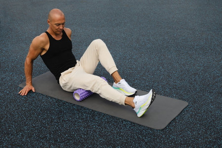 Athlete man exercising and stretching legs on foam roller on sports ground. Outdoor activity....