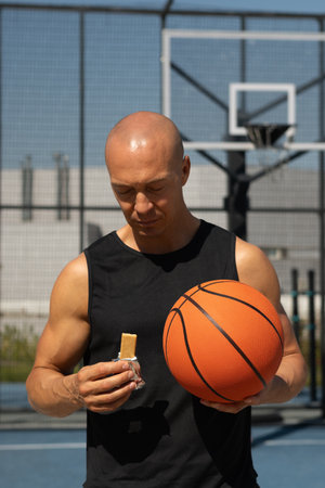 Portrait of basketball player man eating energy bar. Muscular athlete snacks on protein bar while...