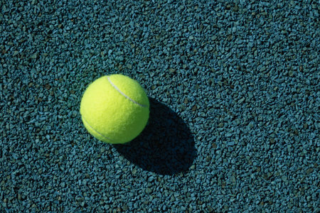 Yellow tennis ball with sunny shadow on blue tennis court with artificial covering. View from above. Summer sports.