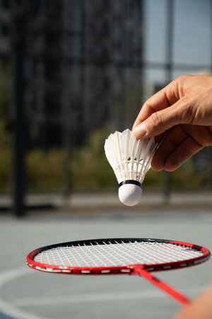 White badminton shuttlecock in man hand and badminton rackets in sunny shadow. Sport badminton court. Outdoors. Game moment. Vertical. Close up.の写真素材