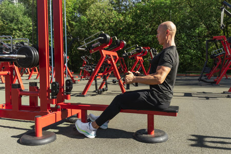 Lat rowing exercises. Adult man training at public gym park. Outdoors activity. Fitness coach show...