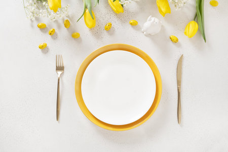 Easter table setting with spring yellow tulips, white plates, bunny, eggs on white background. Happy...