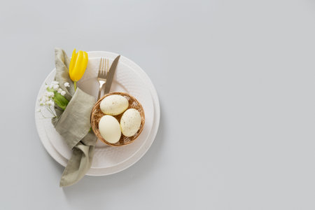Easter table setting with spring yellow tulip, white plates, eggs in nest isolated on gray...