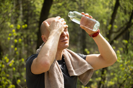 Sporty adult caucasian man refreshing himself water from bottle against forest at summer heat after...