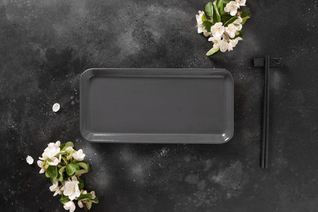 Empty rectangular plate for asian food with black chopsticks on black background with spring blossom...
