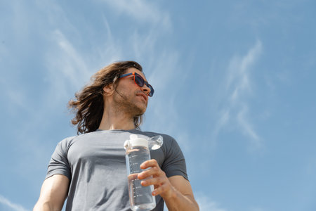 Man in athletic wear with water bottle outdoors against blue sky background. Summer heat weather. Hero view. Thirst. Close up. Banner.の写真素材
