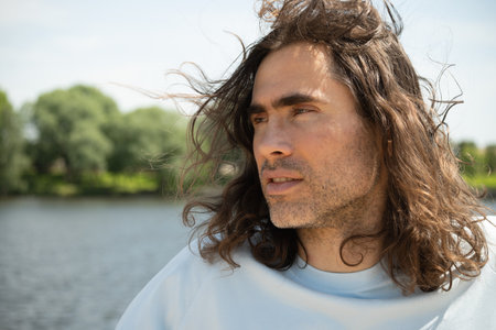 Portrait of thoughtful handsome young caucasian brunette man with long curly hair standing near river. Summer vacation and walk. Slow living. Side viewの写真素材