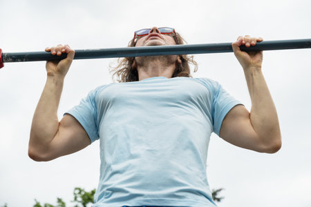 Sporty man wearing blue t-shirt and sunglasses making pull ups monkey bars outdoors and looking up, focused on fitness activity. Summer outdoor setting.の写真素材