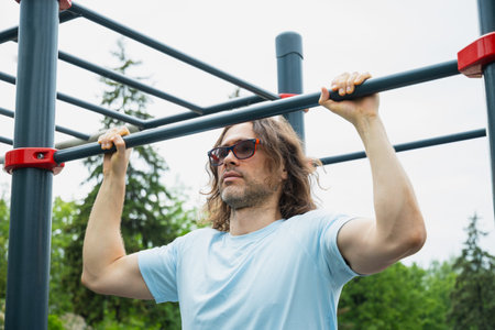 Man wearing sunglasses making pull ups monkey bars outdoors, focused on fitness activity. Sunny...