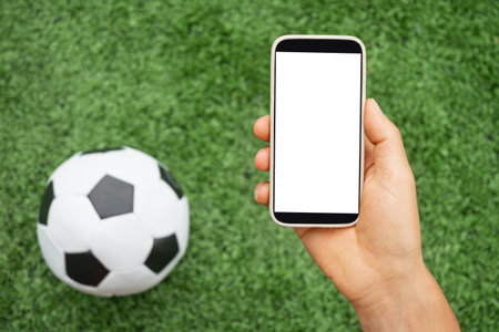 Man hand holds smartphone with white blank screen next to soccer ball on green grass. Online. Modern technology with traditional sports. Space for text. View from above. Empty display .の写真素材