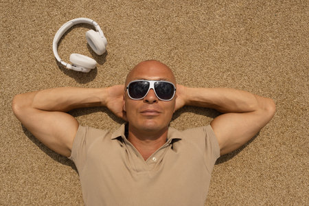 Sun-tanned caucasian man lying down in sunglasses near white headphones on beige background. Relax, enjoy and dream. Slow living. Concept summer vacation.の写真素材